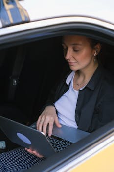 A woman in a black blazer using a laptop inside a car, showcasing remote work lifestyle.